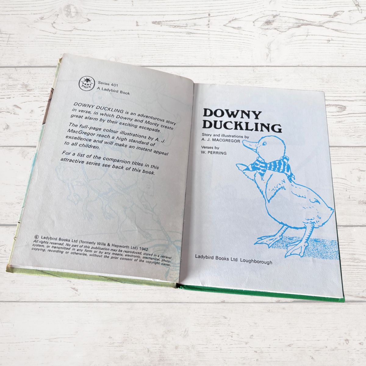 An open children's book titled 'Downy Duckling' from Ladybird Books Ltd, showing the back cover with text and the front cover with a blue line drawing of a duckling, placed on a white wood surface.