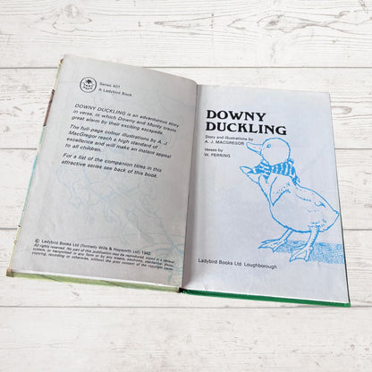 An open children's book titled 'Downy Duckling' from Ladybird Books Ltd, showing the back cover with text and the front cover with a blue line drawing of a duckling, placed on a white wood surface.