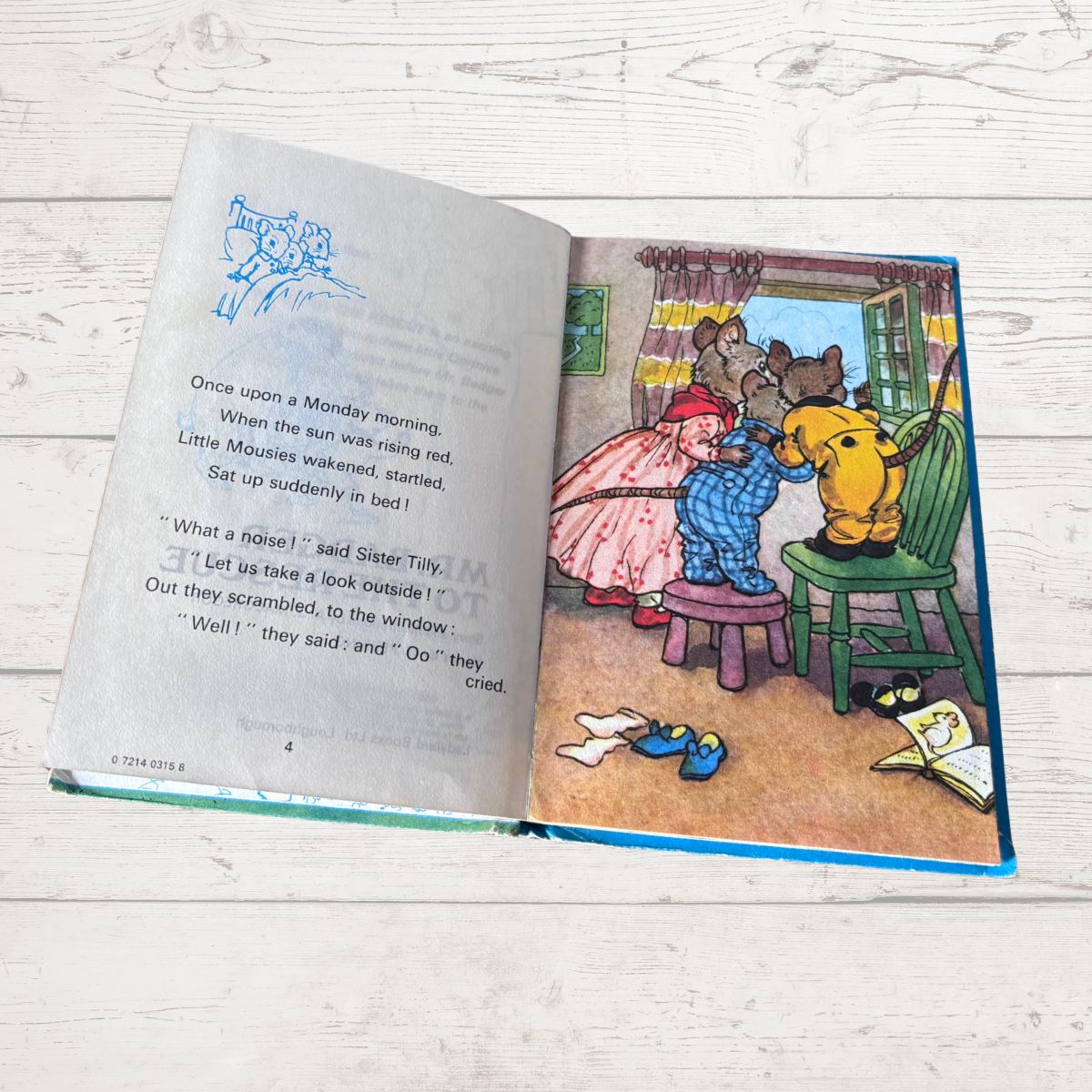 An open children's book laid flat on a light wood surface, showing printed text on the left page and a colorful illustration on the right page with cartoon animals in a room.