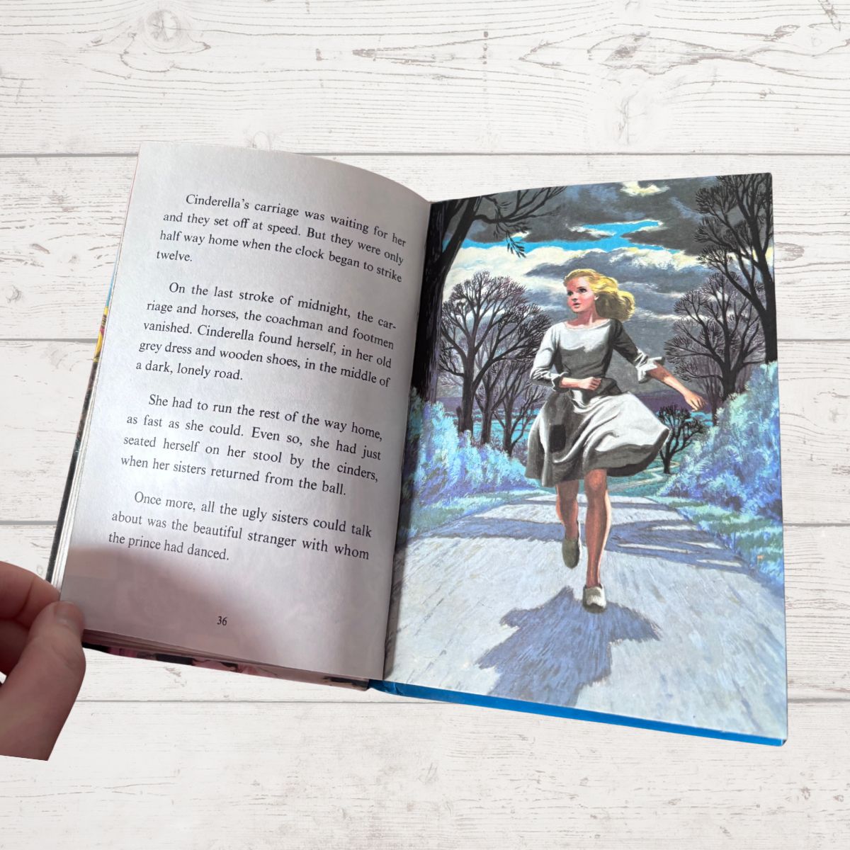 An open children's book showing printed text on the left page and a colorful illustration of a girl running through a snowy landscape on the right page, photographed on a light wood surface with a hand holding the left edge.