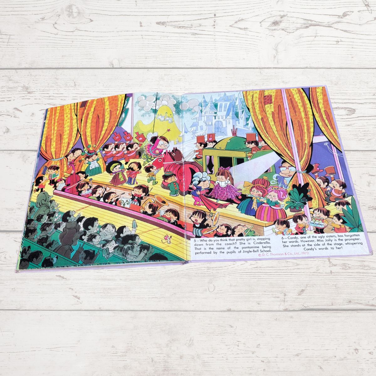 A colorful printed book or comic page laid flat on a light wood surface, showing an illustrated scene with many cartoon characters, large orange curtains, and text blocks along the bottom.