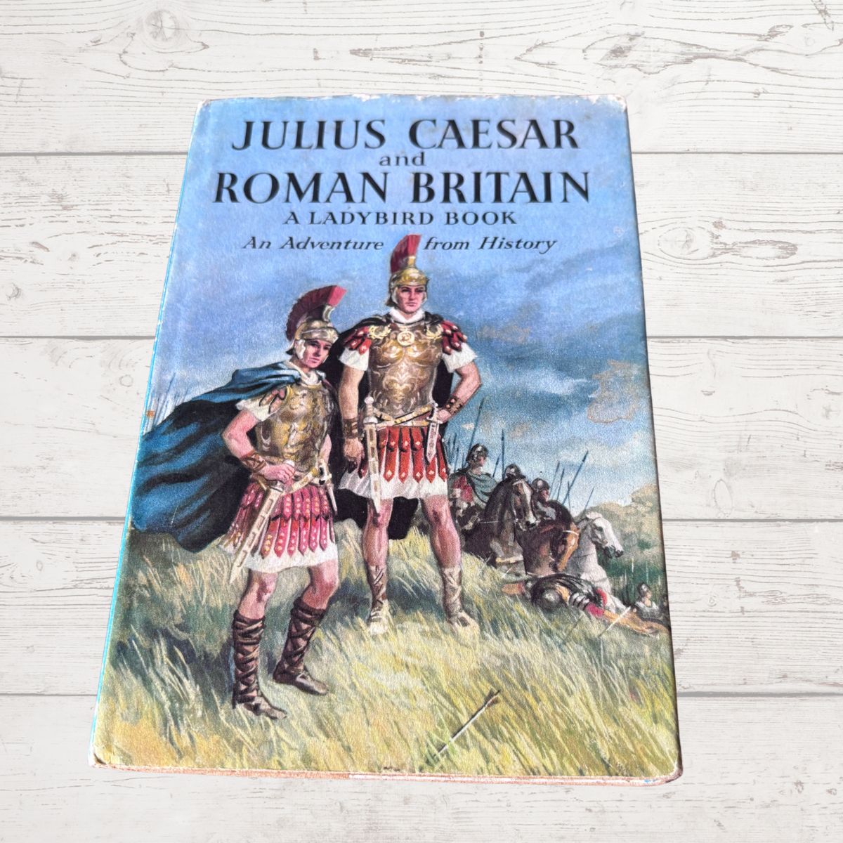 A vintage hardcover book titled 'Julius Caesar and Roman Britain: A Ladybird Book' lying flat on a white wooden surface. The cover features a colorful illustration of Roman soldiers in armor standing in a grassy field under a blue sky.