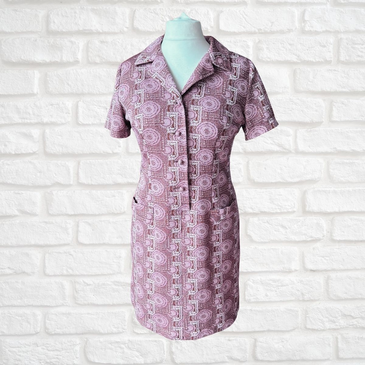A short-sleeve collared button-front dress displayed on a mannequin torso against a white painted brick wall. The fabric has a repeating geometric pattern in dusty pink, mauve, and white.