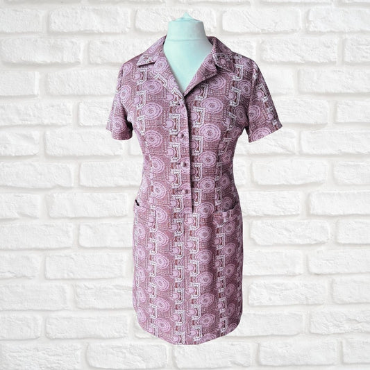 A short-sleeve collared button-front dress displayed on a mannequin torso against a white painted brick wall. The fabric has a repeating geometric pattern in dusty pink, mauve, and white.