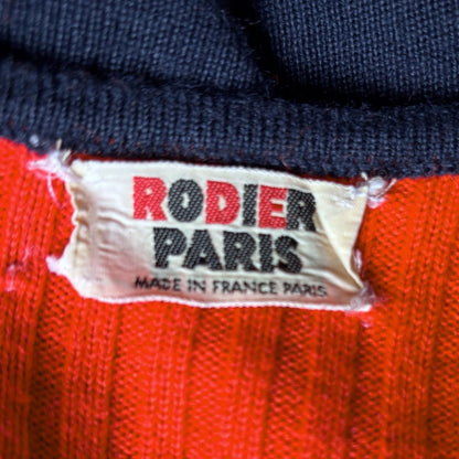 Close-up of a knit garment label reading 'RODIER PARIS' in red and black on a cream tag, with dark navy ribbed knit at the top and bright red knit at the bottom.