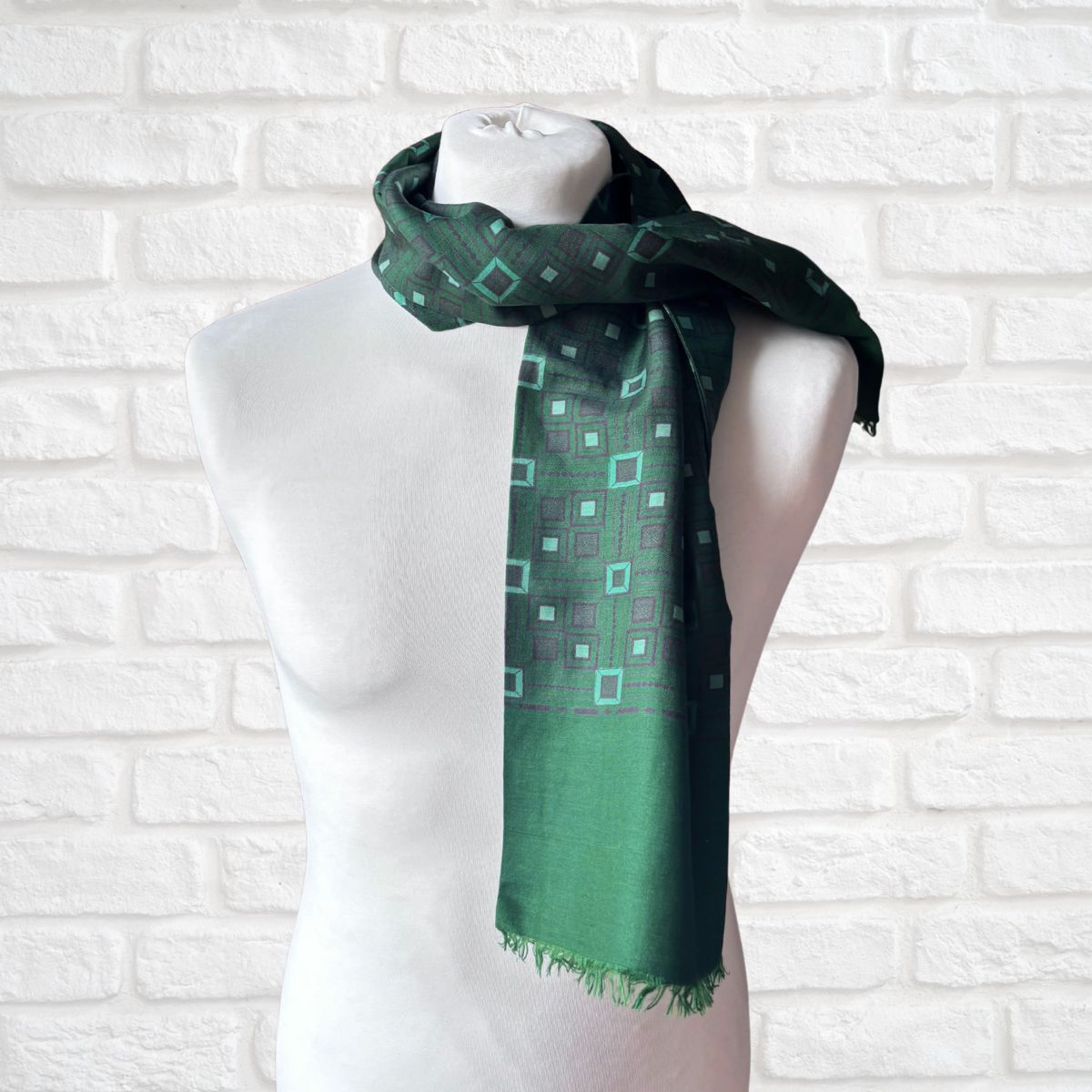 A dark green scarf with a geometric diamond pattern in light green and purple, draped on a white mannequin torso against a white brick wall background.