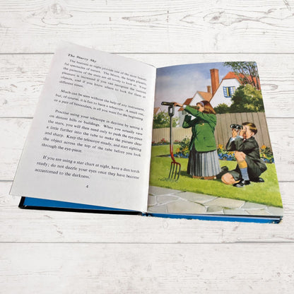 An open children's book lying flat on a white wood surface, showing a printed story page with text on the left and an illustrated scene on the right of a woman in a garden using a telescope.