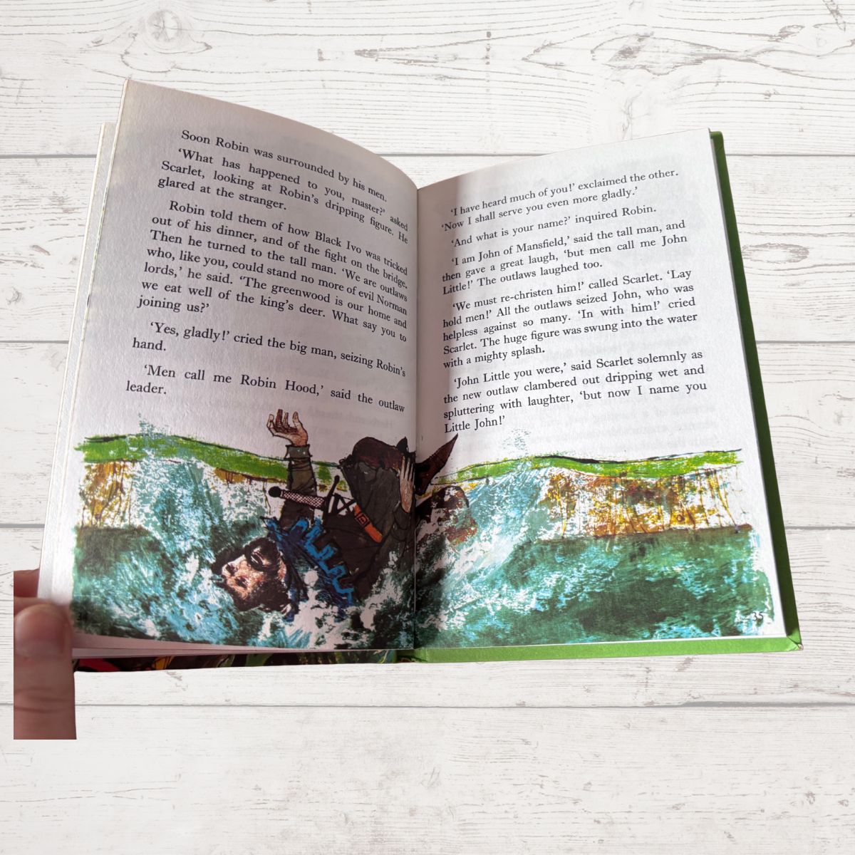 An open children's book lying on a white wood surface, showing printed text on both pages and colorful illustrated scenes along the bottom. No person or animal is visible.