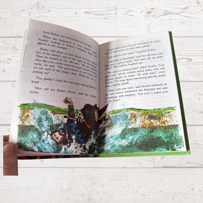 An open children's book lying on a white wood surface, showing printed text on both pages and colorful illustrated scenes along the bottom. No person or animal is visible.