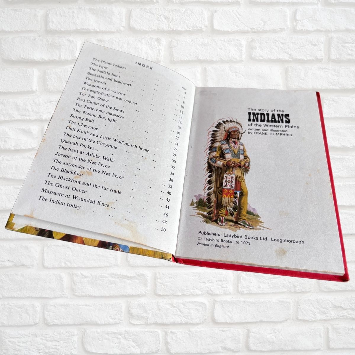 An open vintage hardcover book titled 'The story of the Indians' lying on a white brick background. The left page shows a table of contents, and the right page features an illustration of an Indigenous warrior.