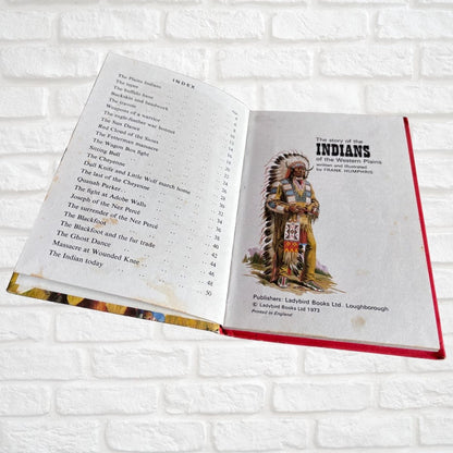 An open vintage hardcover book titled 'The story of the Indians' lying on a white brick background. The left page shows a table of contents, and the right page features an illustration of an Indigenous warrior.