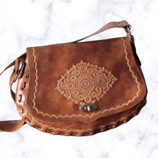 A brown leather shoulder bag with an embossed mandala pattern on the front flap, decorative stitching, and a metal clasp, shown on a white marble background.