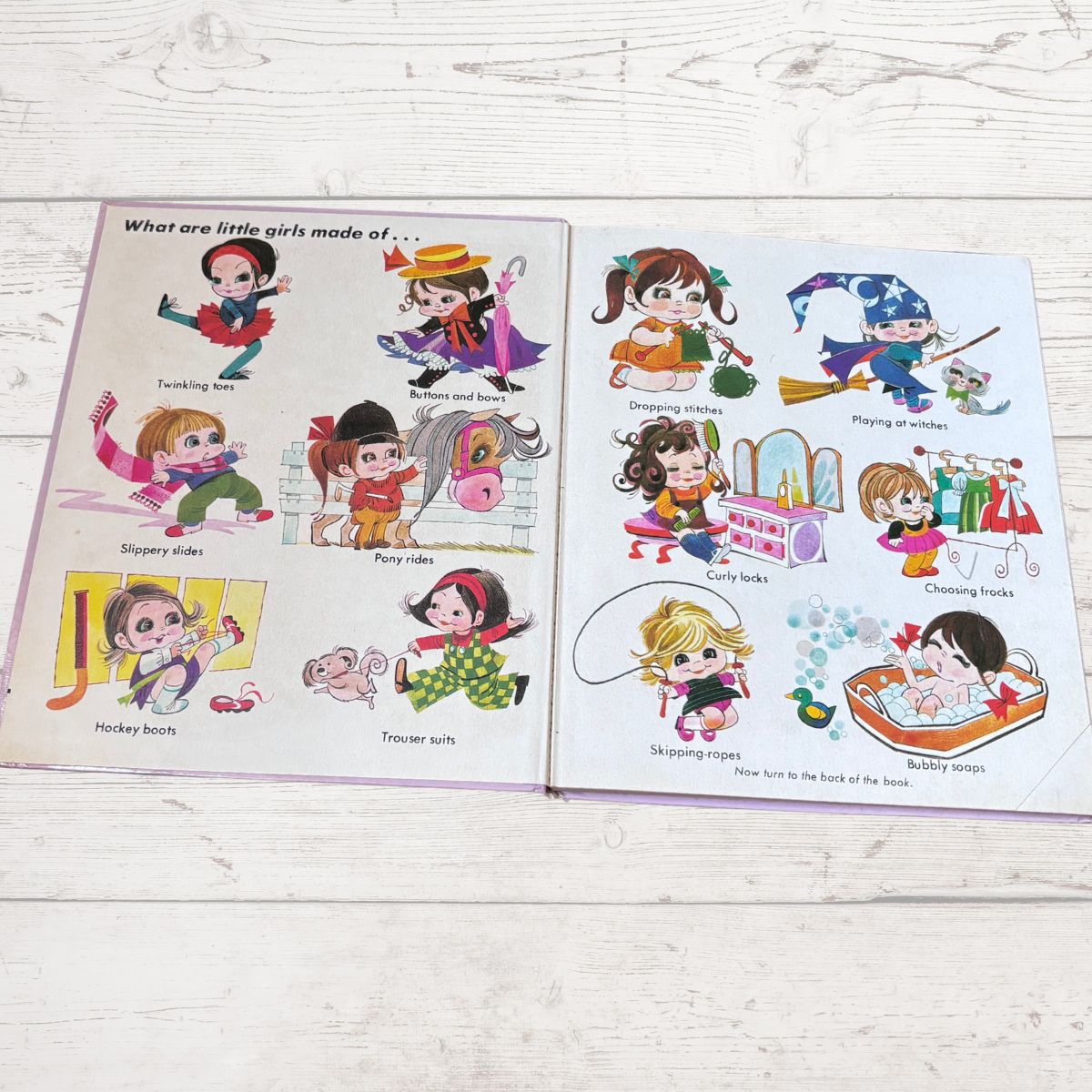 An open children's  book showing cartoon illustrations of little girls with labels such as 'Fancying hats,' 'Pony rides,' 'Hockey boots,' and 'Trouser tails,' placed on a light wood surface.