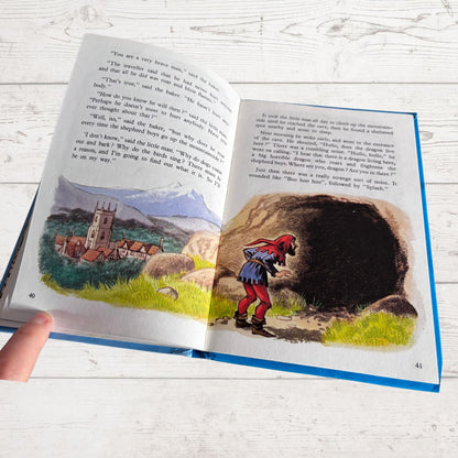 An open children's picture book lying on a white wood surface, showing two illustrated pages with text. A