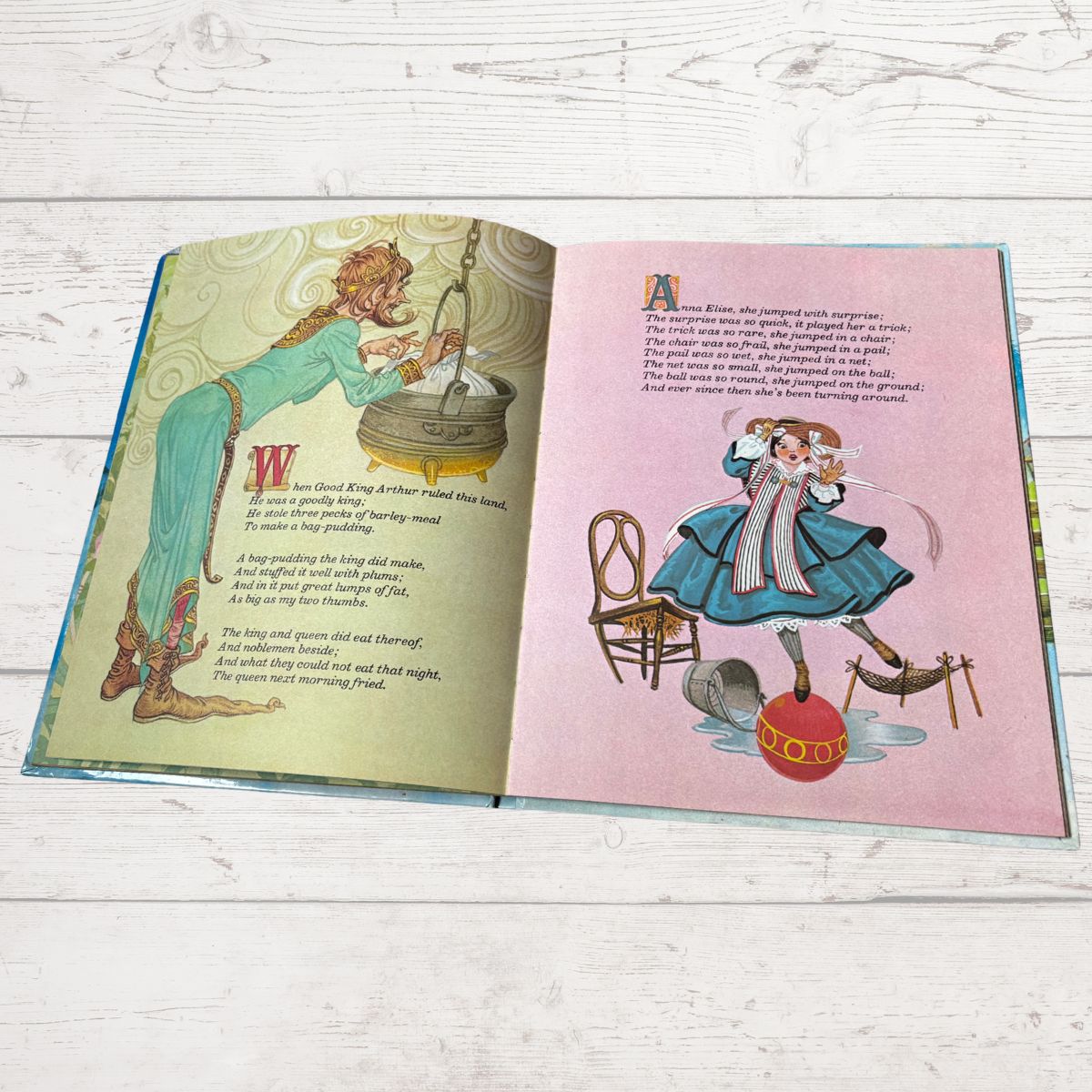 An open children's picture book spread on a whitewashed wood surface, showing illustrated pages with text and colorful drawings.