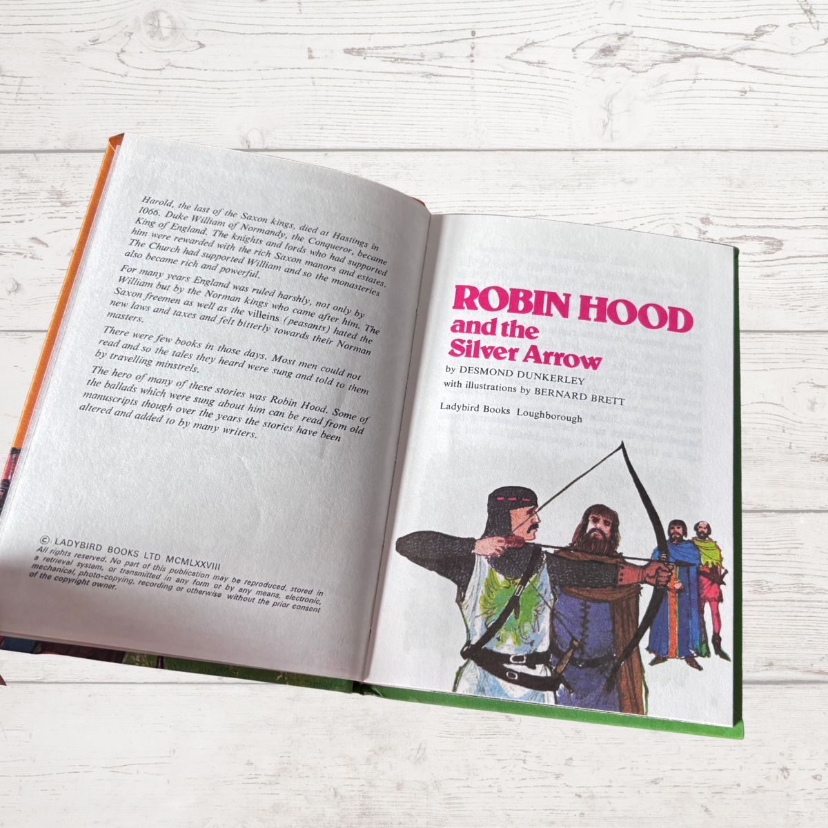 An open children's book titled 'Robin Hood and the Silver Arrow' laid flat on a white wood surface. The left page contains black text, and the right page has a colorful illustration of Robin Hood with an arrow and other figures, with the title in large pink text.