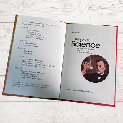 An open hardcover book titled 'The story of Science, Book 2' laid flat on a white wood surface. The left page contains a list of recommended books, and the right page features the title, author text, and a circular portrait illustration of a man holding a glass of water.