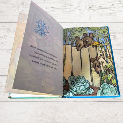 An open children's book lying on a white wood surface, showing a illustrated page with three mice behind a wooden fence and text on the left page.
