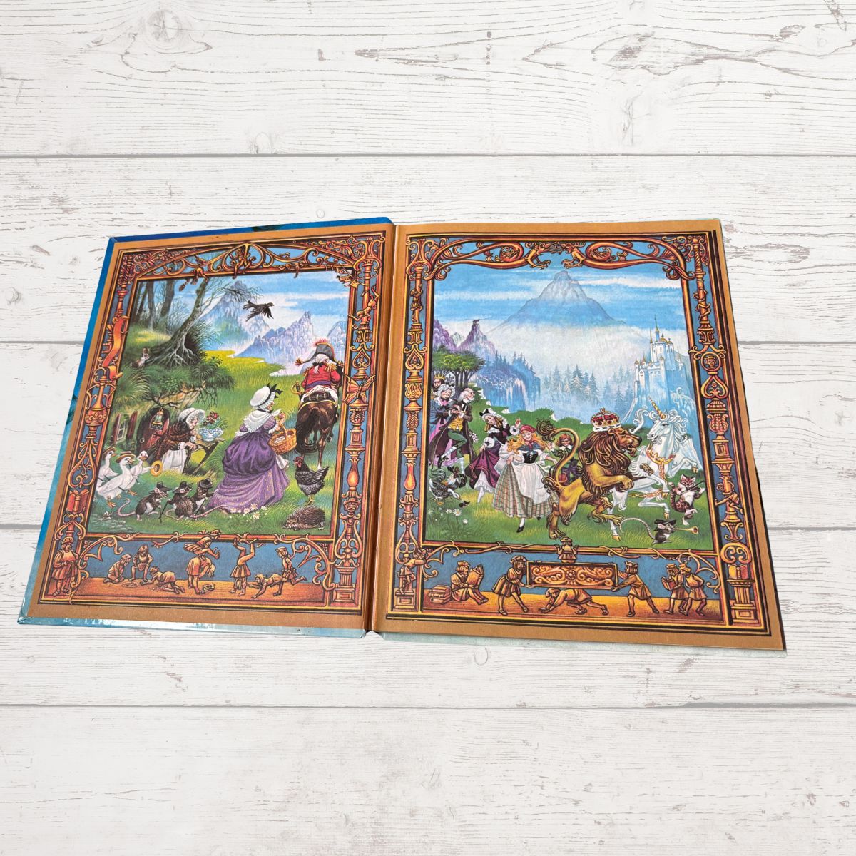 Two vintage illustrated book pages with ornate borders, displayed side by side on a white wood plank surface. The left card shows a forest scene with figures and animals, and the right card shows a mountainous landscape with people and horses.
