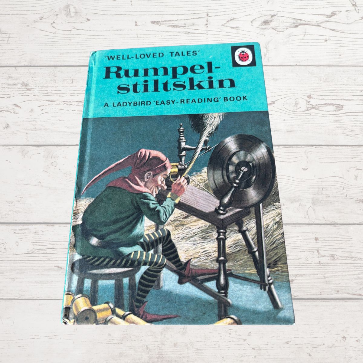 A vintage hardcover children's book titled 'Rumpelstilskin' from Well-Loved Tales, featuring a turquoise top band and an illustration of a fairy spinning wool on a wheel. The book is photographed on a light whitewashed wood surface.
