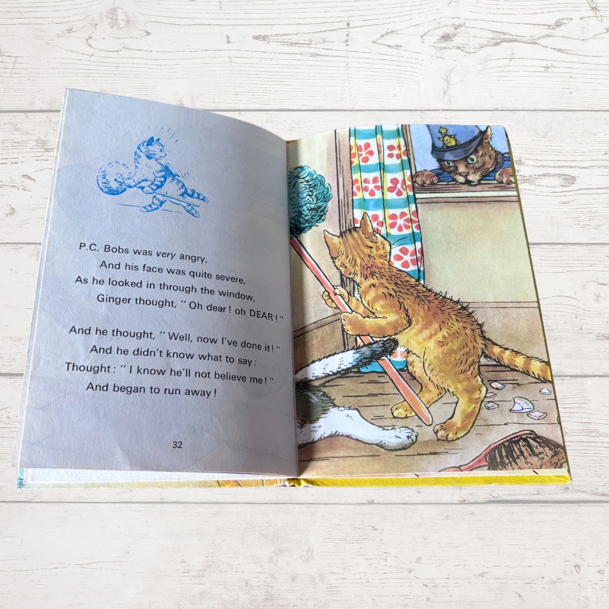 An open children's book lying on a light wood surface, showing a printed story with text on the left page and an illustration of an orange cat chasing a mouse on the right page.