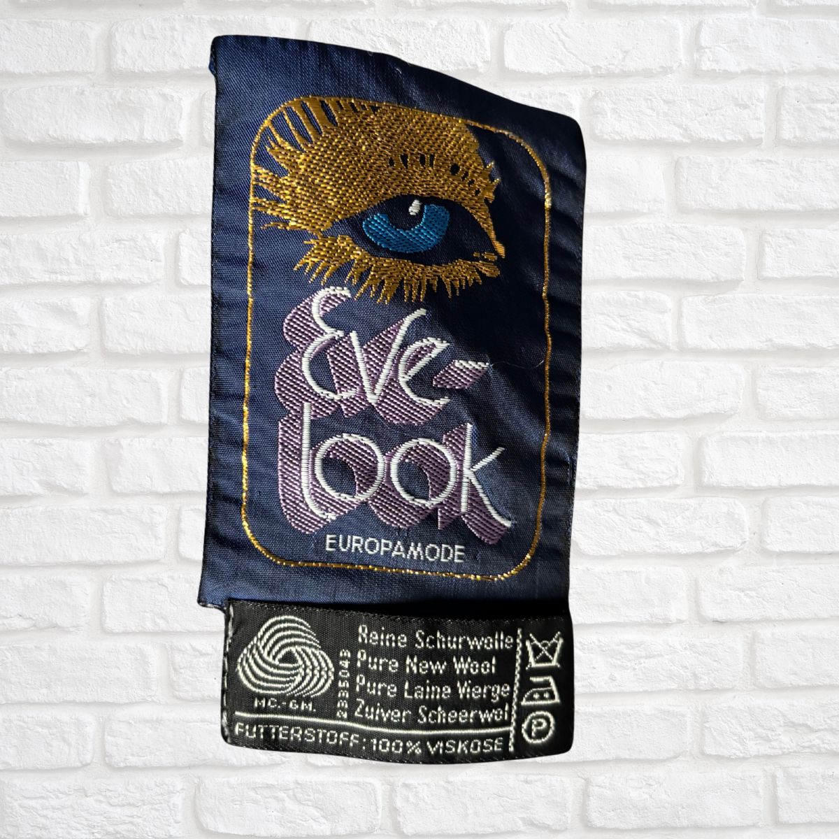 Embroidered label with eye design and text on a white brick wall background