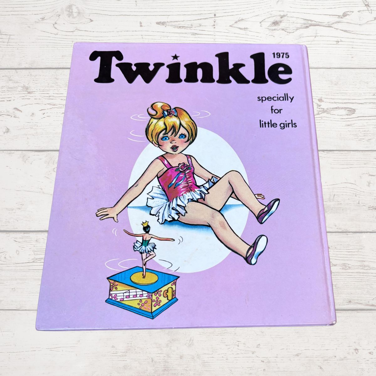 A vintage 1975 children's book titled 'Twinkle' for little girls, featuring a lavender cover with an illustration of a ballerina and a music box, placed on a light wood background.