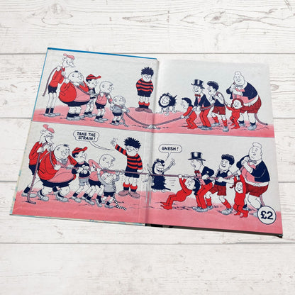 An open vintage comic  book lying flat on a white wood surface. The pages show a hand-drawn cartoon scene with multiple characters in red, navy, black, and white, plus a small 'S2' mark in the lower right corner.
