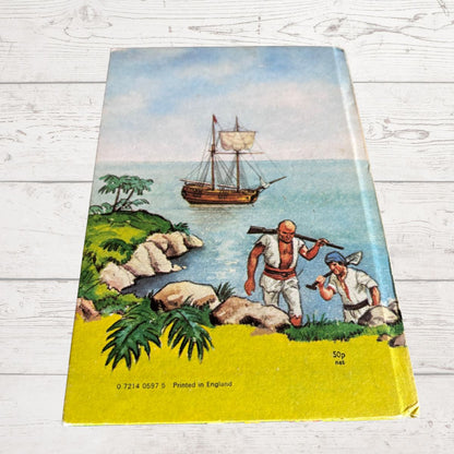 Vintage illustrated book  showing a coastal scene with a sailing ship, rocky shoreline, palm trees, and two figures near the water, photographed on a white wood surface.
