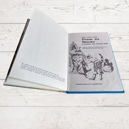 An open children's book titled 'Puss in Boots' laid flat on a light whitewashed wood surface. The pages are white with black printed text and an illustration, and the book has a blue edge visible along the open cover.