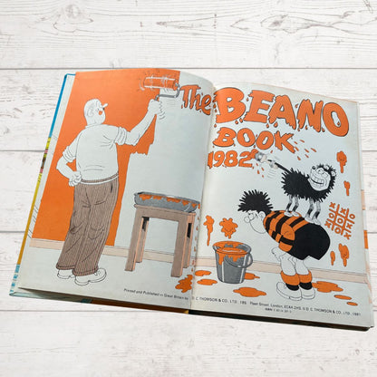 Open book showing the cover and a page from 'The Beano Book 1982' on a wooden surface.