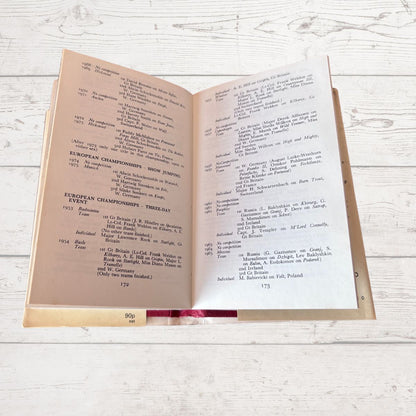 An open vintage book showing printed tables and text, photographed on a light whitewashed wooden surface.