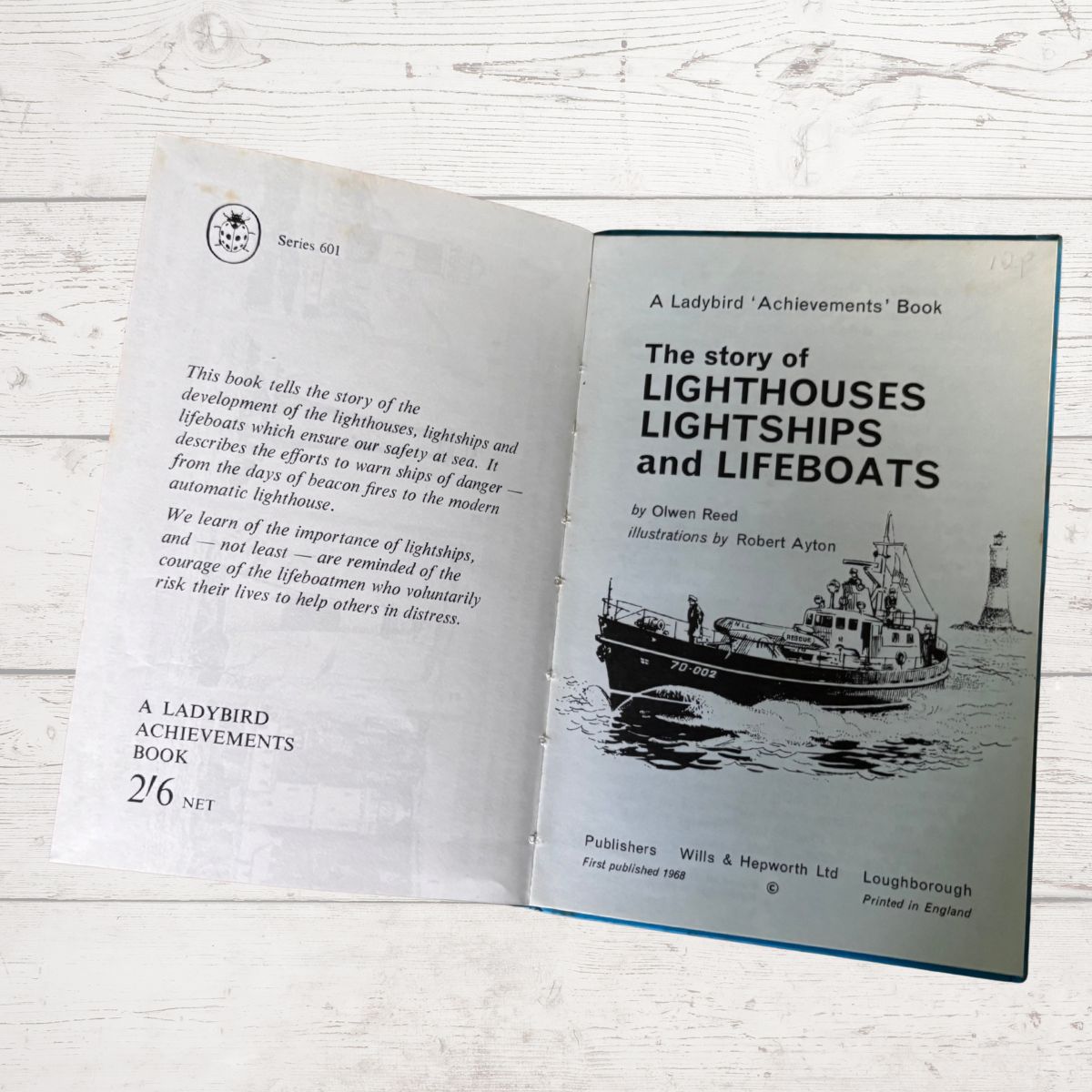 An open children's book titled 'The story of LIGHTHOUSESHIPS AND LIFEBOATS' from the 'A Ladybird Achievements' series, showing the front cover with a black-and-white ship illustration and the back cover with text, photographed on a white wood surface