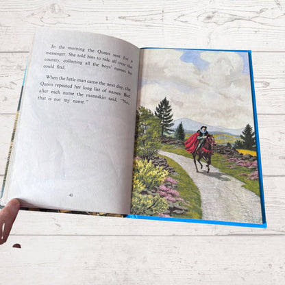 An open children's book lying on a white wood surface, showing printed text on the left page and a colorful landscape illustration on the right page. A hand is visible holding the lower left corner of the book.