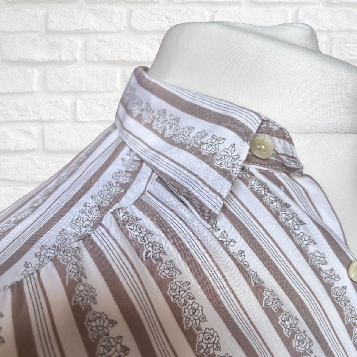 A close-up of a striped and floral-patterned button-up shirt displayed on a white mannequin torso against a white brick wall background.