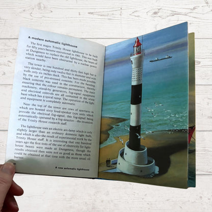 An open vintage book showing printed text on one page and a color photograph of a lighthouse on the other, held by a hand against a light wood background.