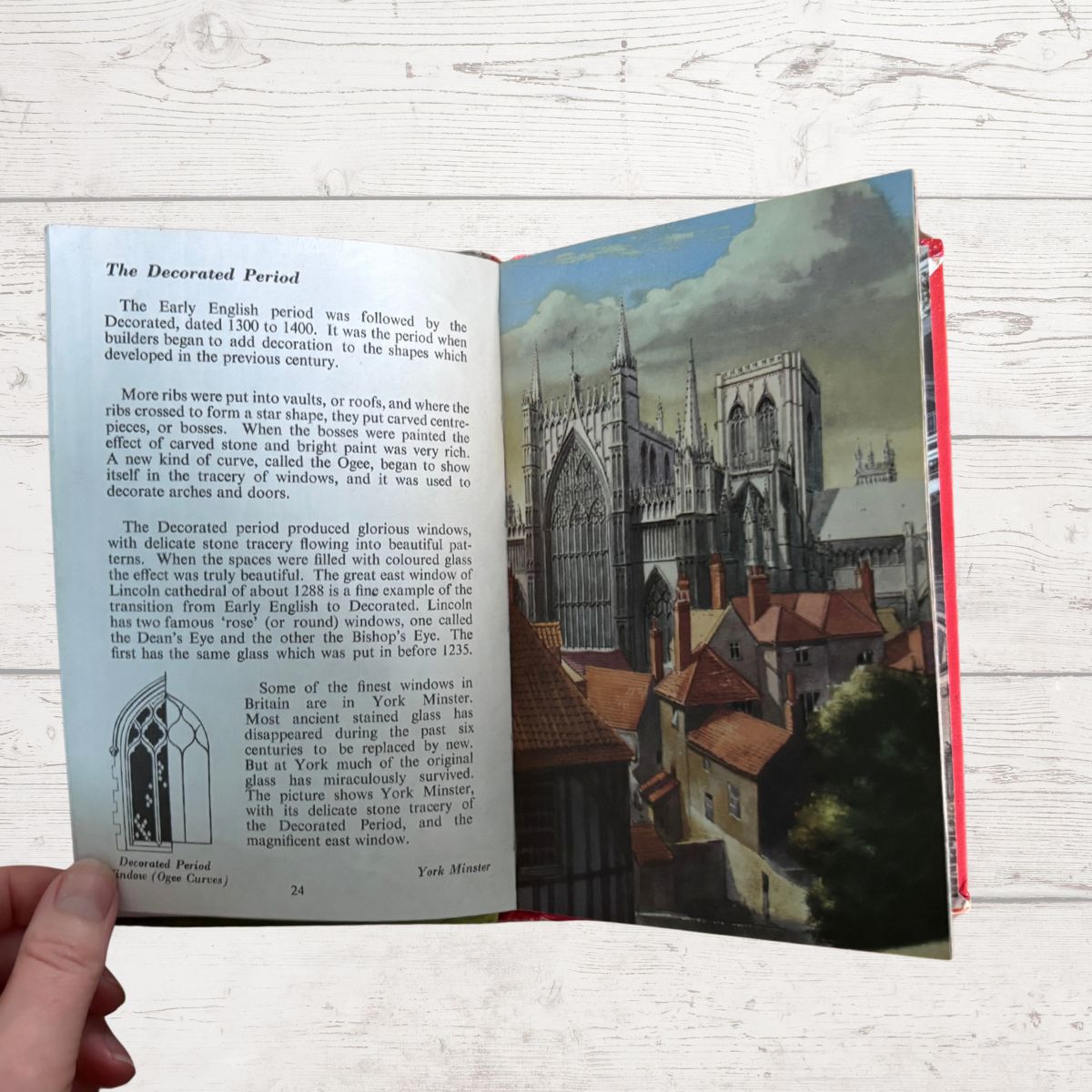An open vintage book showing a page about York Minster's Decorated Period, with a hand holding the left page against a whitewashed wooden background.