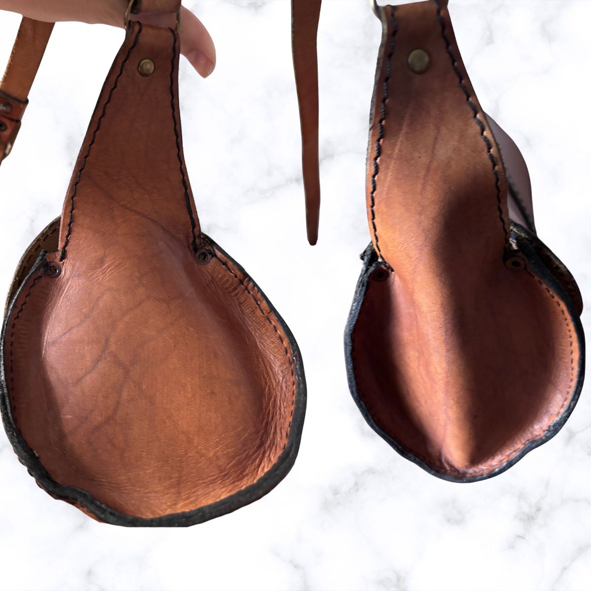 brown leather bag ends shown from the underside on a white marble background, with a hand holding the strap of the left pad.