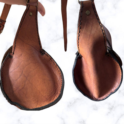 brown leather bag ends shown from the underside on a white marble background, with a hand holding the strap of the left pad.