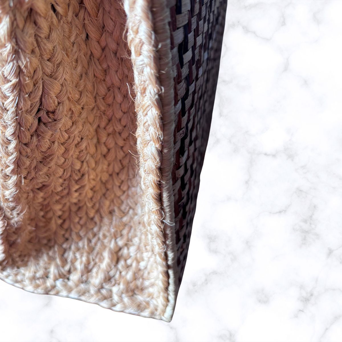 Close-up of a woven basket or bag corner showing a natural beige braided texture and a dark brown patterned weave, set against a white marble background.