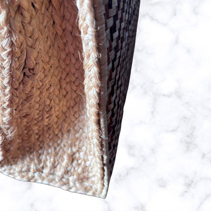 Close-up of a woven basket or bag corner showing a natural beige braided texture and a dark brown patterned weave, set against a white marble background.