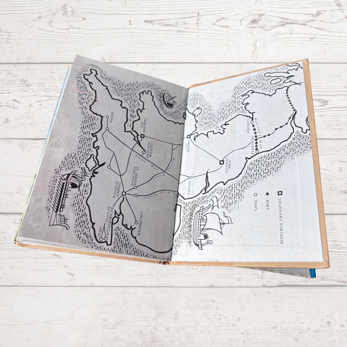 An open illustrated book  lying on a white wood surface, showing two map pages with black line drawings, labels, and small illustrations of ships and buildings.