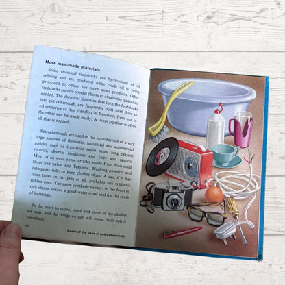 An open children's educational book showing a page about man-made materials and petrochemicals, with a colorful illustration of household items like a washing machine, lamp, and tools. A hand is holding the book against a light wood background.