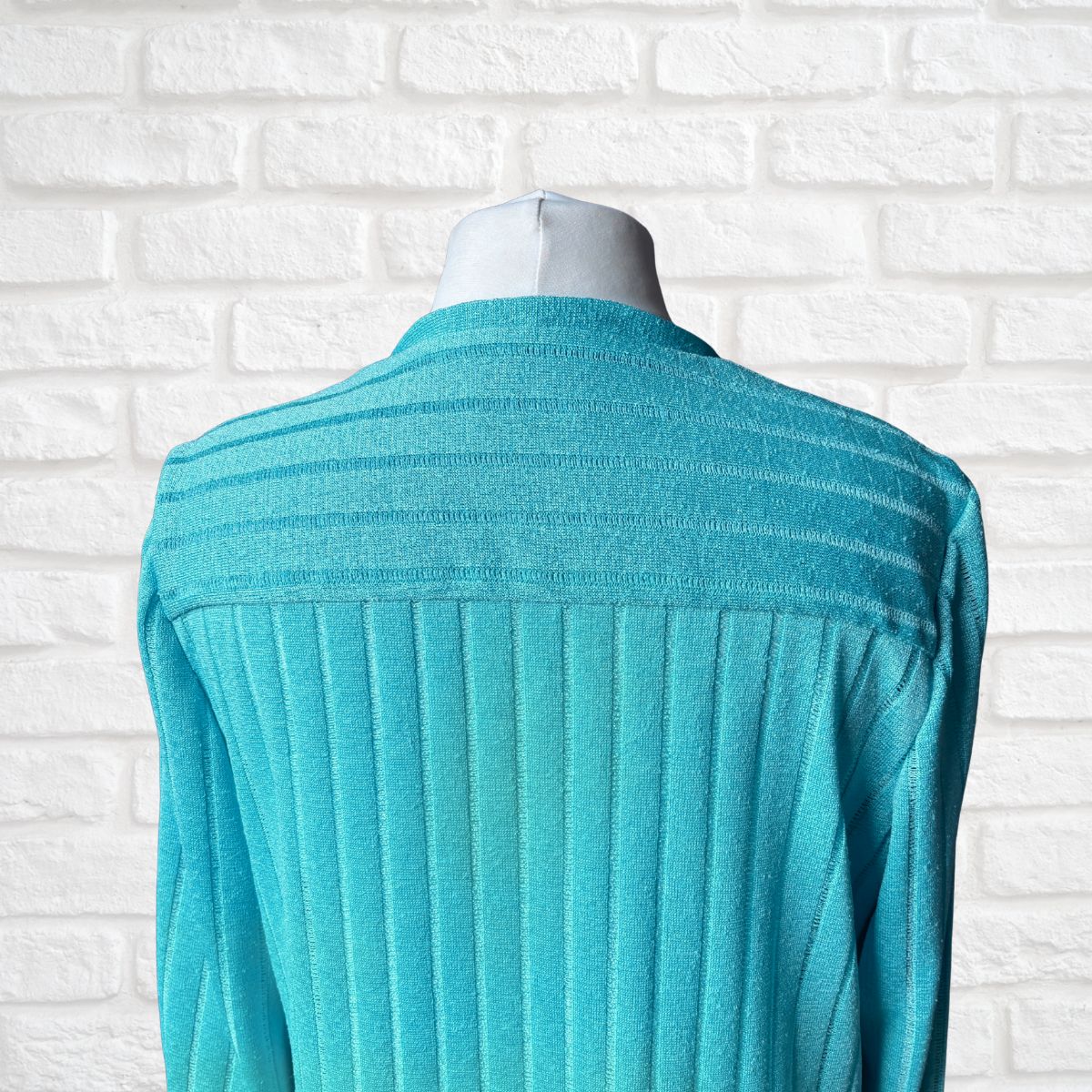 A turquoise ribbed knit cardigan displayed on a mannequin torso against a white brick wall background.