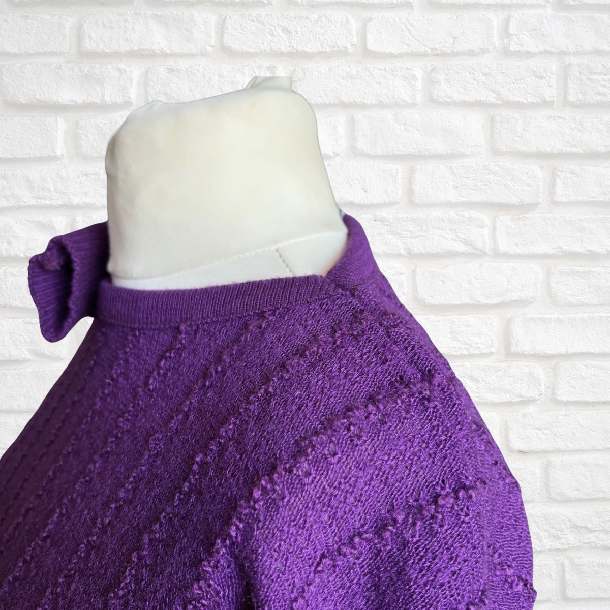 A close-up of a purple knit sweater displayed on a white mannequin torso against a white brick wall background.