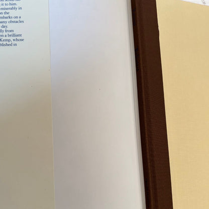 A close-up of a book showing a cream page, a dark brown spine or binding edge, and a light beige textured cover or backing.