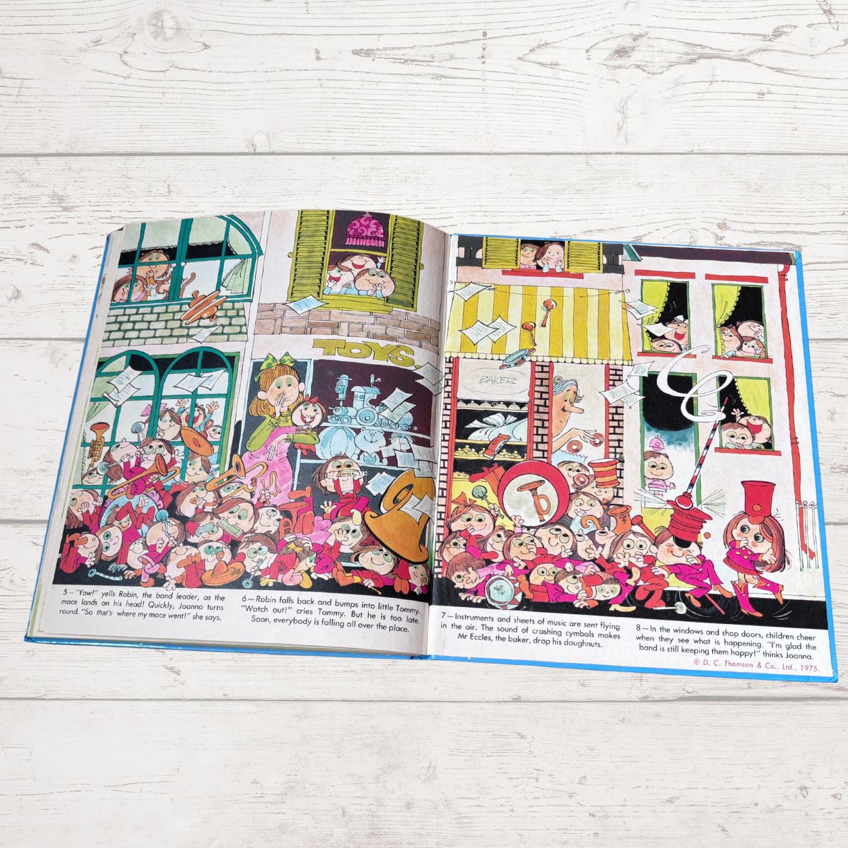 An open children's picture book spread on a light whitewashed wood surface, showing colorful cartoon illustrations and short text on both pages.