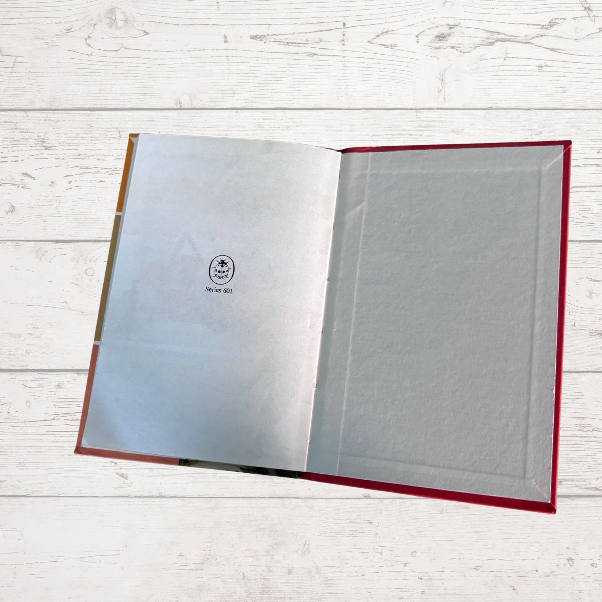 An open book with a red hardcover, laid flat on a white wood-plank surface. The interior pages are white