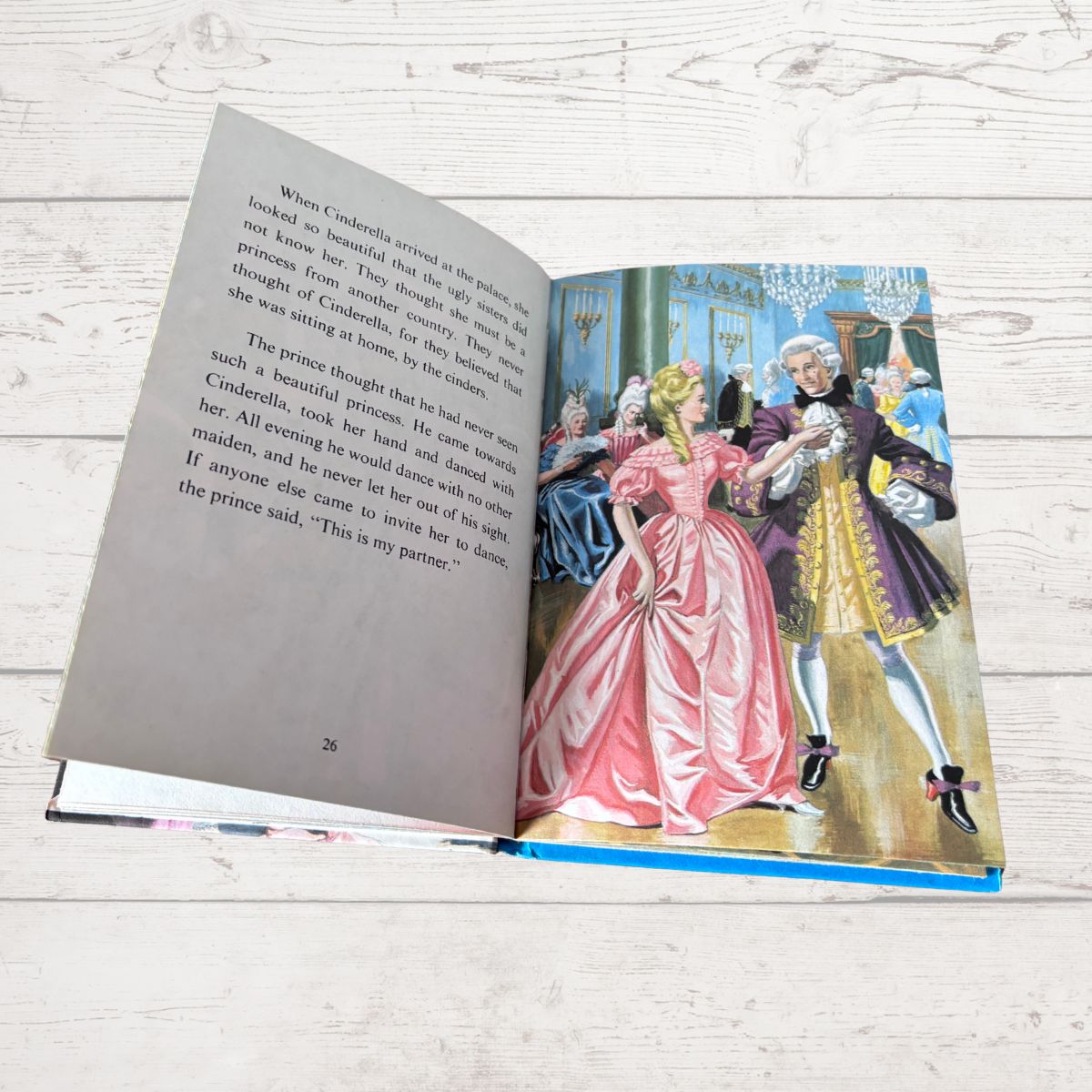 An open children's book showing a printed story page with text on the left and a colorful illustration of Cinderella and the prince on the right, placed on a light wood surface.