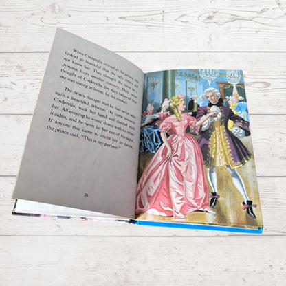 An open children's book showing a printed story page with text on the left and a colorful illustration of Cinderella and the prince on the right, placed on a light wood surface.